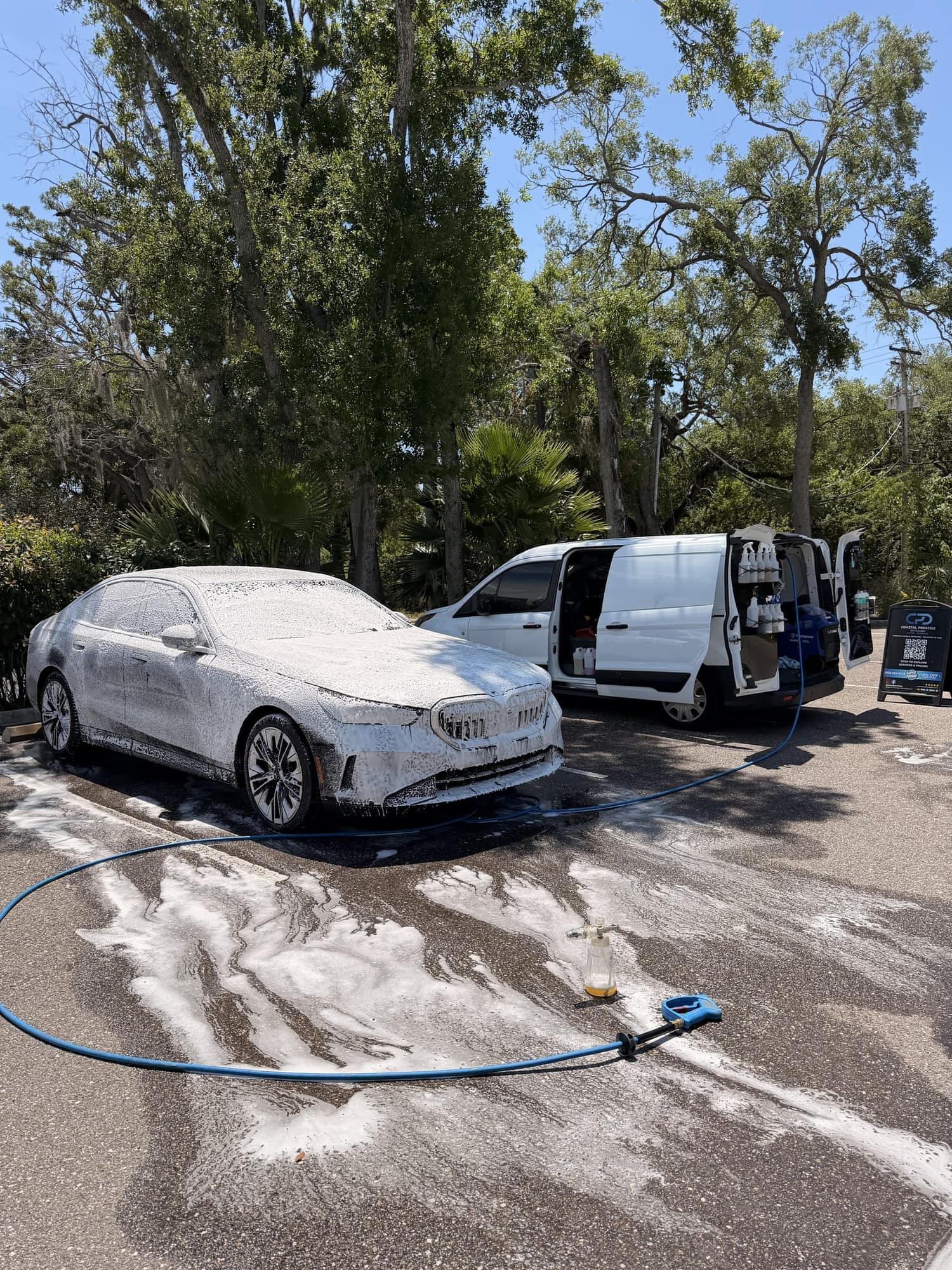 BMW sedan covered in foam on a mobile detail in Wesley Chapel, with the Coastal Prestige Detailing van open alongside and the foam cannon on the ground
