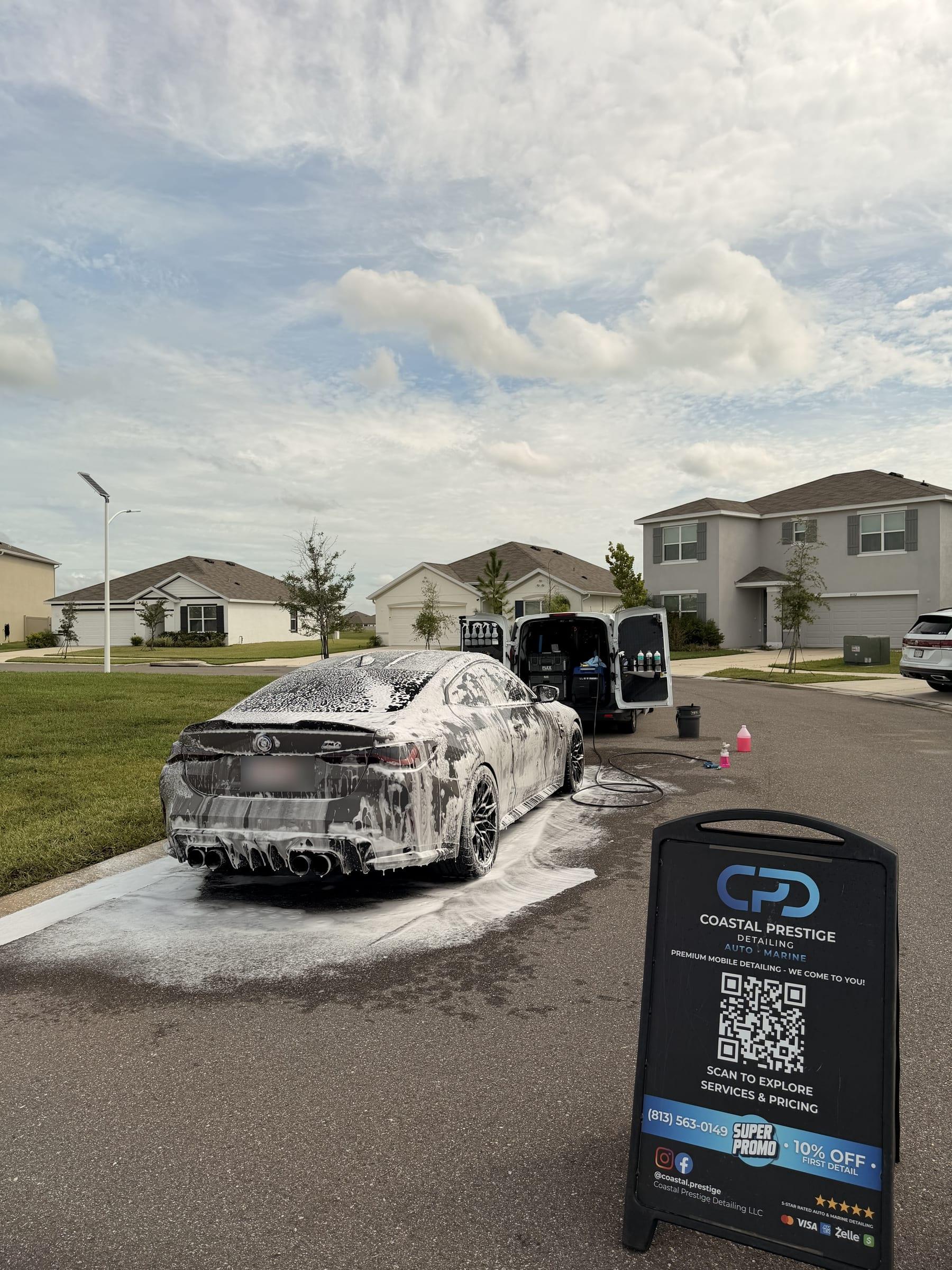 BMW M4 covered in foam on a residential driveway in Wesley Chapel, with the Coastal Prestige Detailing van and sign alongside